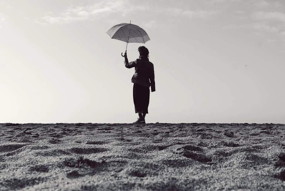 Black and white silhouette of a woman holding an umbrella while standing on a serene beach.