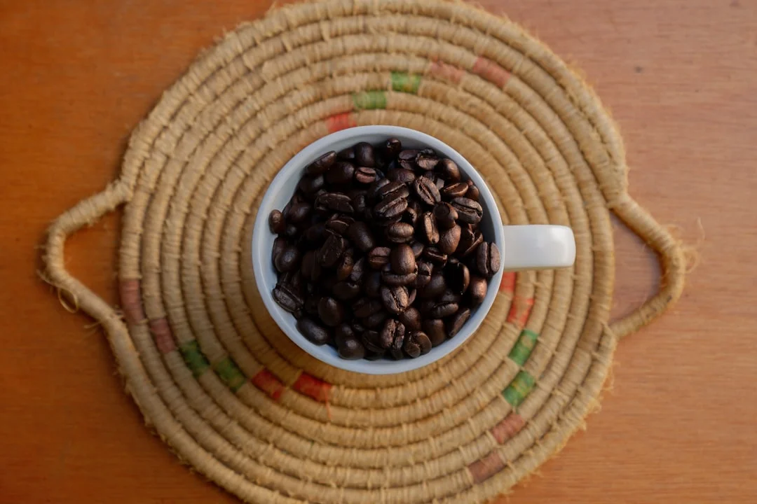 Coffee beans fill a white cup on a woven mat.